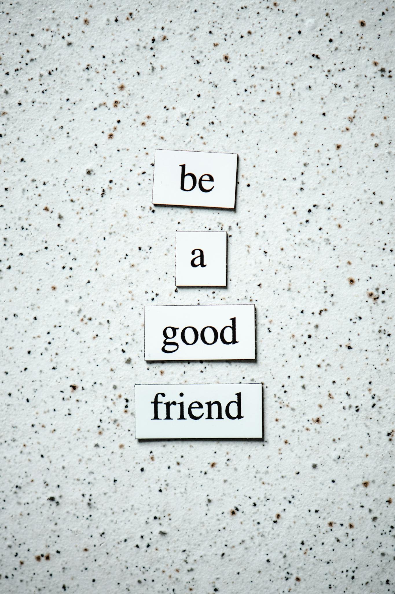 Text tiles 'be a good friend' on a pebbled surface for inspiring friendship.